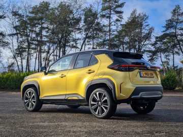 Een gele en zwarte Toyota Yaris Cross compacte SUV staat geparkeerd op een buitenterrein met op de achtergrond bomen en een bewolkte lucht. De auto heeft een Nederlands kenteken.