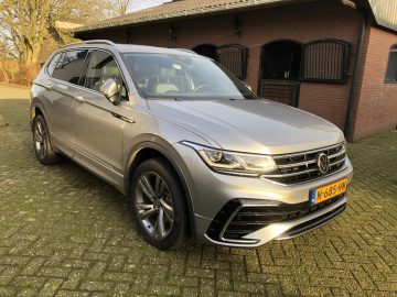 Een metallic zilveren Volkswagen Tiguan SUV staat geparkeerd op een stenen oprit voor een bakstenen gebouw met donkerhouten deuren. Het voertuig heeft een Nederlands kenteken met de vermelding "N-685-HV".
