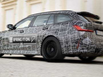 Een gecamoufleerde M3 Touring-stationwagen met verschillende lijnpatronen en de woorden "BRING IT ON" op de zijkant staat geparkeerd op een geplaveide ondergrond voor een crèmekleurig gebouw.