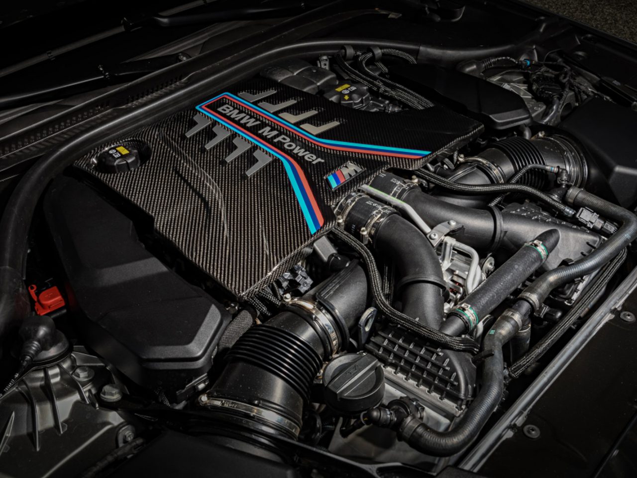 Close-up van een BMW M Power-motor, met ingewikkelde BMW-componenten en koolstofvezeldetails onder de motorkap. Motoronderdelen, kabels en diverse mechanismen zijn duidelijk zichtbaar.