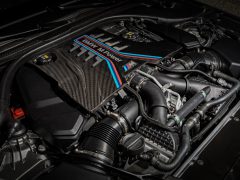 Close-up van een BMW M Power-motor, met ingewikkelde BMW-componenten en koolstofvezeldetails onder de motorkap. Motoronderdelen, kabels en diverse mechanismen zijn duidelijk zichtbaar.