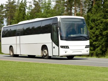 Op een weg met een bosgebied op de achtergrond rijdt een witte tourbus, op de voet gevolgd door een politievoertuig.