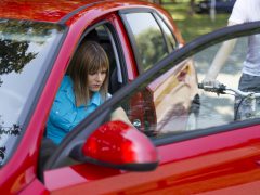 Een vrouw in een blauw shirt zit in een rode auto met de deur open en kijkt naar beneden, terwijl naast haar een automobilist op een fiets voorbijrijdt.