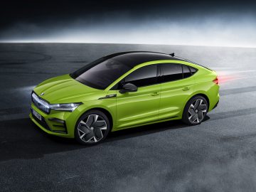 Een groene Skoda Enyaq Coupé staat geparkeerd op de stoep met het achterremlicht verlicht. De auto heeft strakke lijnen, getinte ramen en opvallende lichtmetalen velgen.