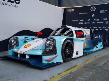 In een binnenomgeving wordt een raceauto op waterstof, ook wel waterstofauto genoemd, tentoongesteld. Er staan logo's van verschillende sponsors, waaronder Shell en Fischer. De auto heeft een strak ontwerp met een combinatie van witte, blauwe en zwarte kleuren.