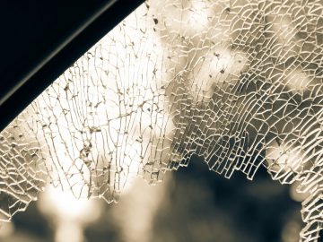 Close-up van een autoruit met gebroken glas, waardoor het zicht naar buiten zichtbaar wordt door talloze scheuren en gebroken segmenten. De achtergrond lijkt wazig, waardoor de autoschade haarscherp wordt benadrukt.