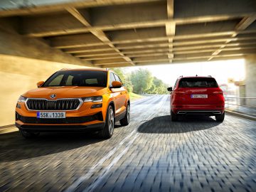 Twee auto's, een oranje SUV en een rode Skoda Karoq, rijden zij aan zij op een geplaveide weg onder een betonnen brug, met zonlicht en groen op de achtergrond.