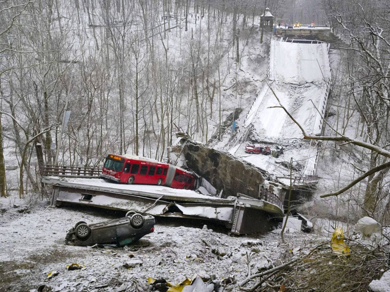 Een met sneeuw bedekte brug is ingestort, waarbij een rode bus en verschillende voertuigen op het puin zijn gestrand. De scène, emblematisch voor de uitdagingen van de Amerikaanse infrastructuur, is omgeven door een bosrijk gebied en op de achtergrond zijn hulpvoertuigen zichtbaar.