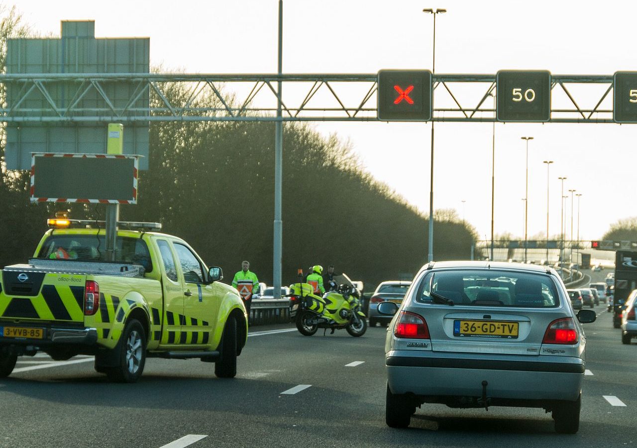 Snelwegscène met voertuigen, waaronder een gele bedrijfswagen en motorfiets. Een bord boven het hoofd toont een 'rode X' en een snelheidslimiet van 50, wat aangeeft dat er een afgesloten rijstrook is. Het negeren van dit signaal kan een fikse boete op de snelweg tot gevolg hebben.