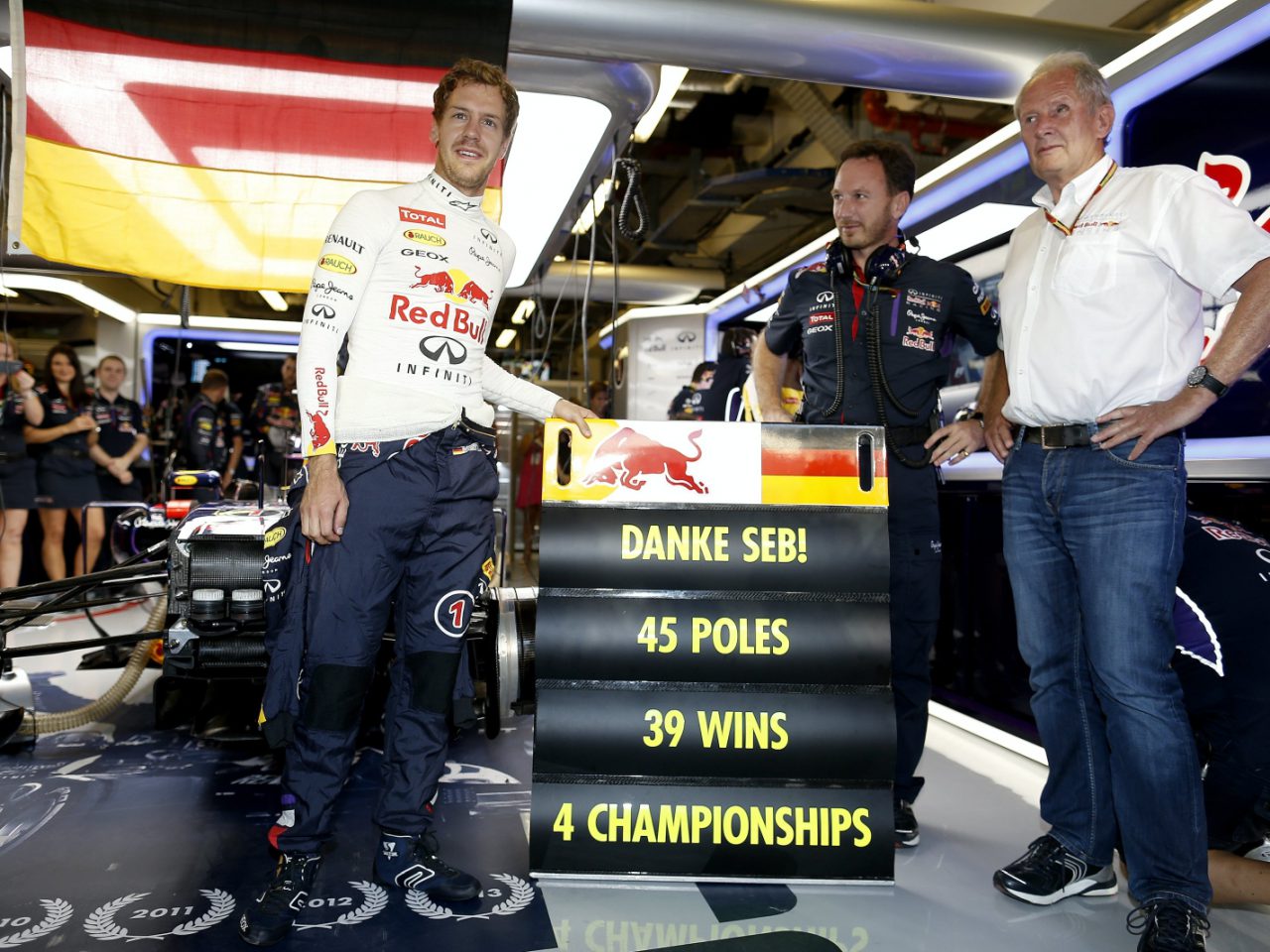 Een autocoureur poseert met drie mannen bij een bord in een racegarage met de tekst "DANKE SEB! 45 POLES, 39 WINS, 4 CHAMPIONSHIPS", met zijn Formule 1-auto en fans op de achtergrond.