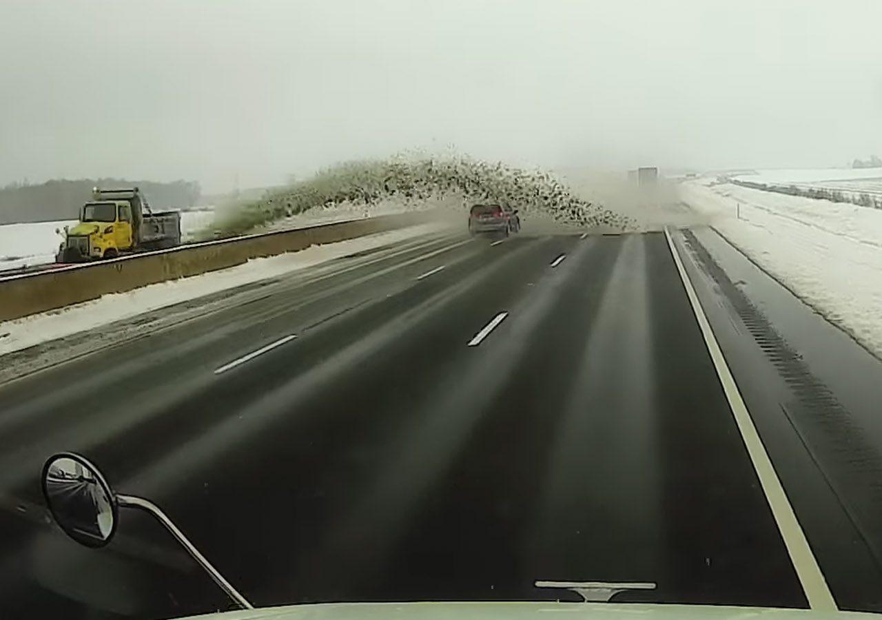 Een sneeuwschuiver spuit een grote hoeveelheid sneeuw en sneeuwbrij op de tegenoverliggende rijstrook van de weg, waardoor passerende voertuigen op een besneeuwde snelweg gedeeltelijk aan het zicht worden onttrokken.