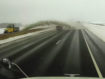 Een sneeuwschuiver spuit een grote hoeveelheid sneeuw en sneeuwbrij op de tegenoverliggende rijstrook van de weg, waardoor passerende voertuigen op een besneeuwde snelweg gedeeltelijk aan het zicht worden onttrokken.