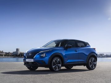 Een blauwe Nissan Juke Hybrid staat geparkeerd op een betonnen ondergrond aan het water, met een helderblauwe lucht boven zijn hoofd en een stadsgezicht op de achtergrond.