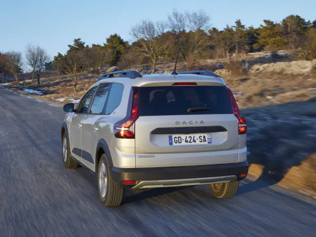 Een witte Dacia Jogger navigeert in de winter over een landelijke weg, terwijl sneeuw de grond en de bomen op de achtergrond bestrooit, wat het decor vormt voor een geïmproviseerde Dacia Jogger-recensie.