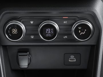 Close-up van het klimaatcontrolepaneel van de Dacia Jogger met drie knoppen, een digitaal temperatuurdisplay ingesteld op 19,5 °C, een USB-poort en een 12V-stopcontact.