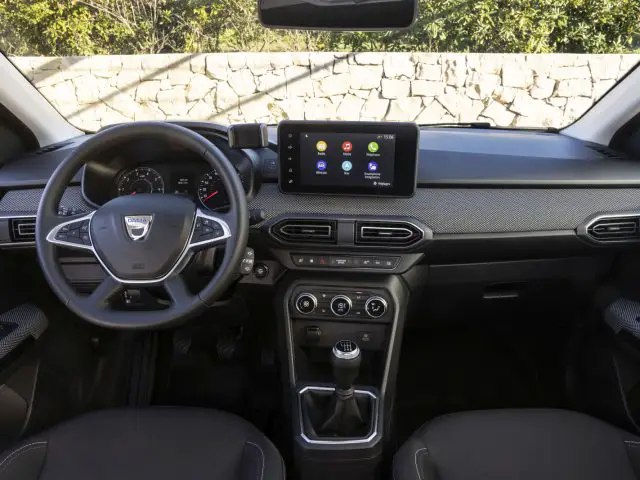 Binnenaanzicht van een Dacia Jogger met het stuur, het dashboard, de versnellingspook en een infotainmentscherm met verschillende apps. De achtergrond is voorzien van een stenen muur en groen gezien door de voorruit.