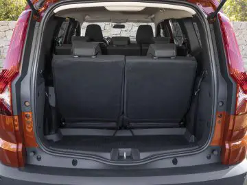 Achteraanzicht van een open, lege kofferbak van een oranje SUV met de achterbank rechtop. Op de achtergrond is een stenen muur zichtbaar, die een typisch voorbeeld is van een typische Dacia Jogger-recensiesetting.