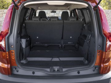 De afbeelding toont de bagageruimte van een oranje Dacia Jogger SUV met de achterbank rechtop. Het interieur is zwart en de kofferruimte oogt ruim en overzichtelijk. Deze Dacia Jogger review benadrukt de indrukwekkende opslagcapaciteit.