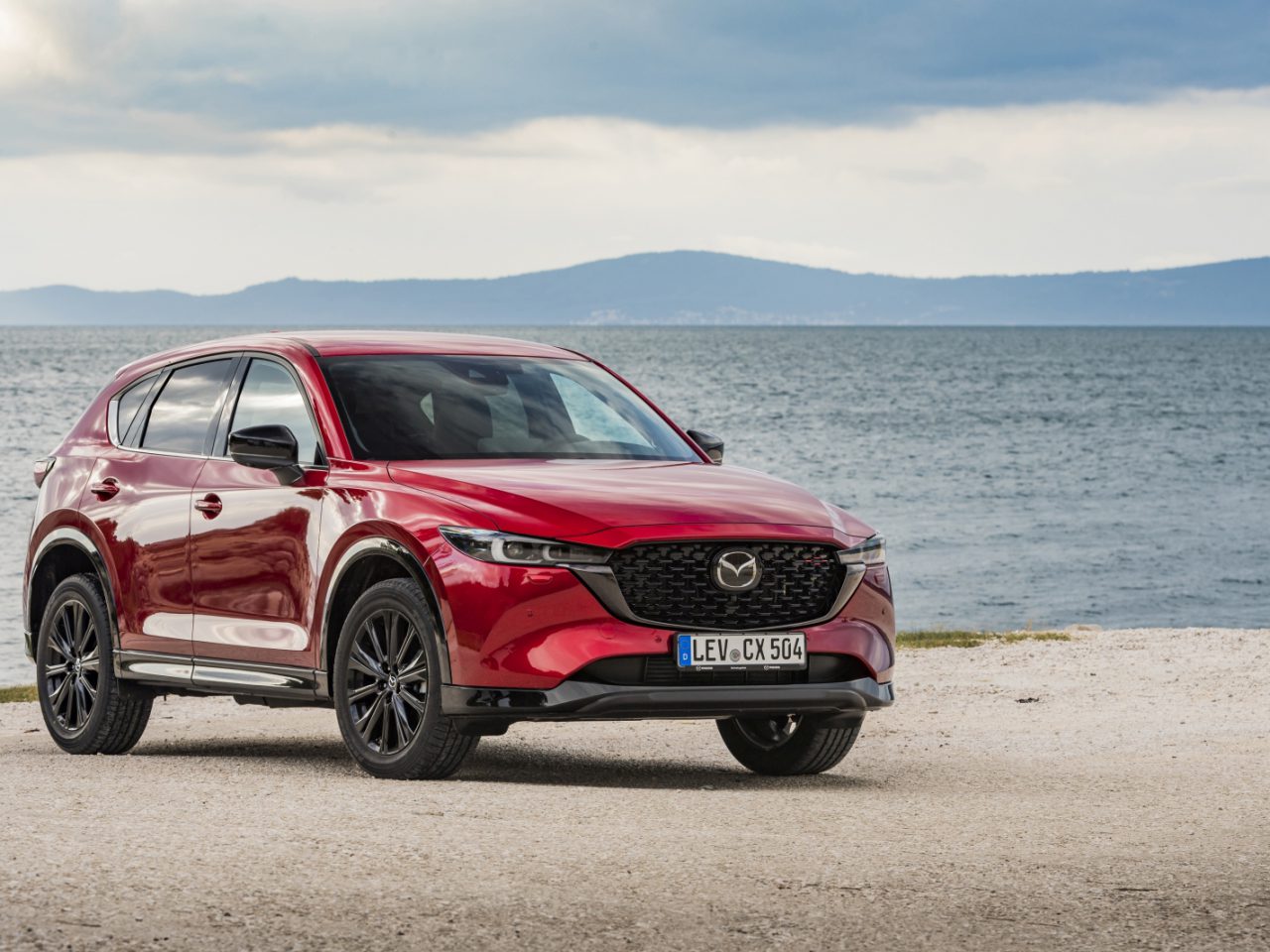 Een rode Mazda CX-5 SUV staat geparkeerd op een zandstrand met een grote watermassa en bergen zichtbaar op de achtergrond onder een bewolkte hemel, wat het decor vormt voor een ideale Mazda CX-5 review.