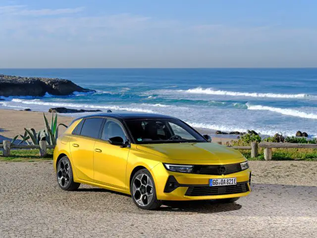 Een gele Opel Astra staat geparkeerd op een kasseistrook met uitzicht op de oceaan en de golven op het strand op de achtergrond. De lucht is helder en blauw.