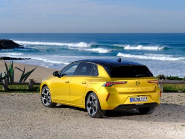 Een strakke geel-zwarte Opel Astra staat geparkeerd op een geplaveid oppervlak aan de kust, met op de achtergrond golven op de kust.