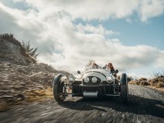 Een vintage Morgan driewielige auto rijdt over een schilderachtige, bochtige weg met heuvels en bewolkte luchten op de achtergrond. Er zitten twee mensen in de auto en genieten van de rit.