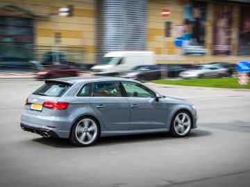Een grijze auto rijdt door een drukke stadsstraat met verschillende andere voertuigen op de achtergrond. Onder hen slingert een gestolen Audi door het verkeer. Op de achtergrond zijn ook gebouwen en een busje zichtbaar.
