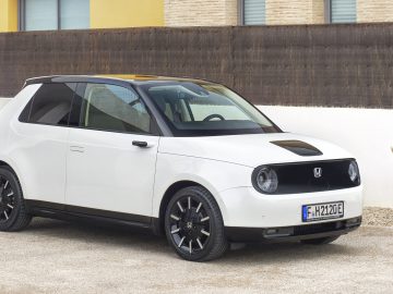 Een witte compacte elektrische auto, perfect om door elke milieuzone te navigeren, staat geparkeerd op een grindoprit. De auto is voorzien van zwarte accenten, een panoramisch dak en ronde koplampen. Een beige muur en enkele potplanten bevinden zich op de achtergrond.