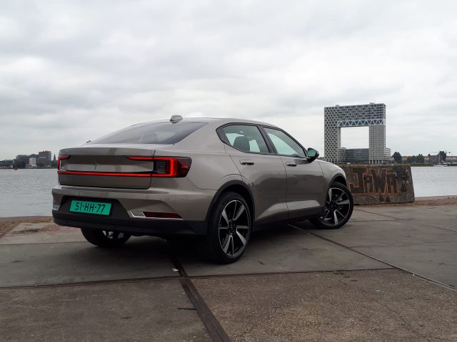 Een grijze elektrische auto geparkeerd op een betonnen ondergrond nabij een waterkant, binnen de milieuzone, met een groot gebouw en een bewolkte lucht op de achtergrond.