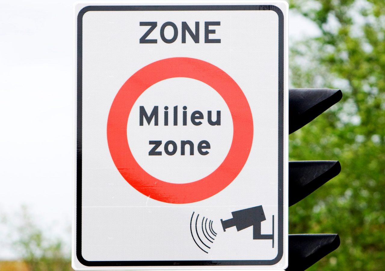 Op een verkeersbord met een rode cirkel staat "ZONE Milieu zone" en staat een afbeelding van een bewakingscamera. Bomen staan op de achtergrond, wat aangeeft dat dit een aangewezen milieuzonegebied is.