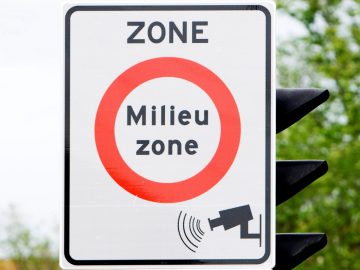 Op een verkeersbord met een rode cirkel staat "ZONE Milieu zone" en staat een afbeelding van een bewakingscamera. Bomen staan op de achtergrond, wat aangeeft dat dit een aangewezen milieuzonegebied is.