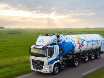 Een waterstofauto met het merk FrieslandCampina en een boodschap over nulemissie rijdt over een landelijke weg met groene velden onder een bewolkte hemel.