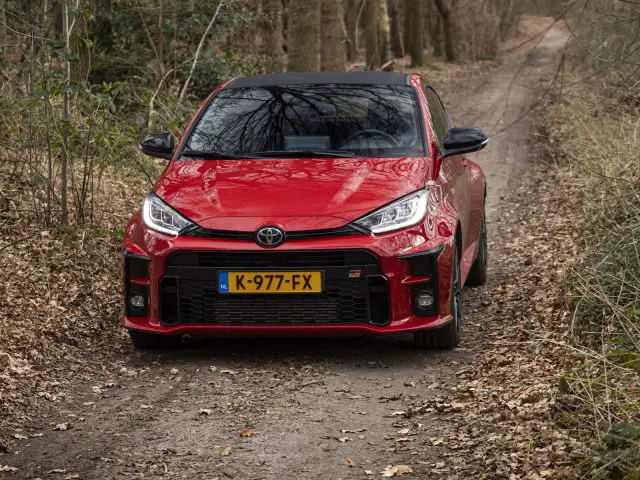 Een rode Toyota GR Yaris, trots voorzien van een Nederlands kenteken, rijdt over een smal, met bomen omzoomd onverhard pad bedekt met droge bladeren en bladeren.