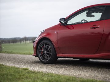 Close-up van de voorkant van een rode Toyota GR Yaris met zwarte velgen, geparkeerd op een landelijke weg, met een grasveld en een bewolkte lucht op de achtergrond.