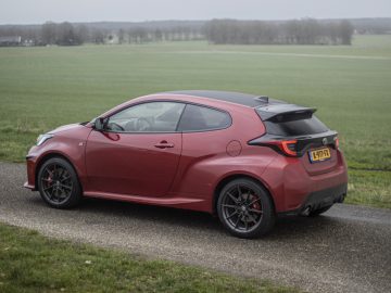 Een rode Toyota GR Yaris hatchback staat geparkeerd op een smalle weg met een groen veld en bewolkte lucht op de achtergrond. Het kenteken van de auto is zichtbaar.