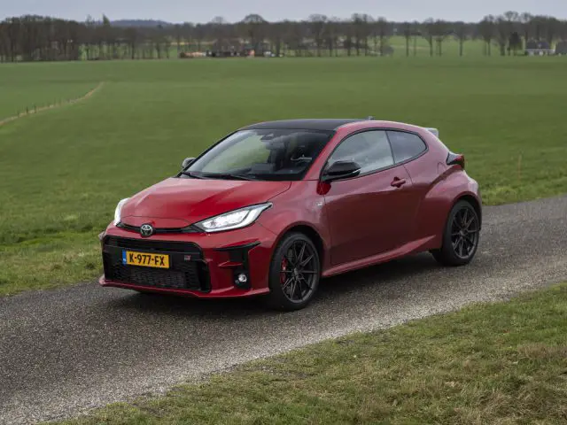 Een rode compacte Toyota GR Yaris met Europees kenteken staat geparkeerd op een landelijke weg, met groene velden en een bewolkte lucht op de achtergrond.