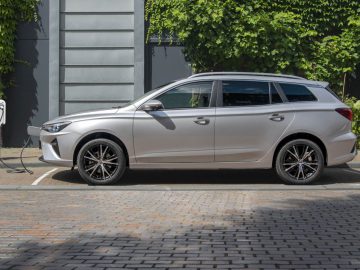 Een zilveren MG5 elektrische auto staat geparkeerd en wordt opgeladen bij een openbaar laadstation naast een gebouw met met klimop begroeide muren.