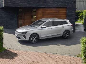 Een zilveren MG5 Electric stationwagen staat geparkeerd op een moderne oprit voor een donker bakstenen huis met een houten garagedeur.