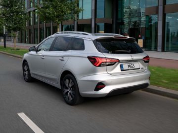 Een slanke zilveren MG5 Electric glijdt moeiteloos over een stedelijke weg, omlijst door weelderige bomen en moderne gebouwen.