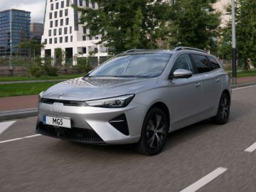 Een zilveren MG5 Electric stationwagen rijdt op een stadsweg met moderne gebouwen en groen op de achtergrond.