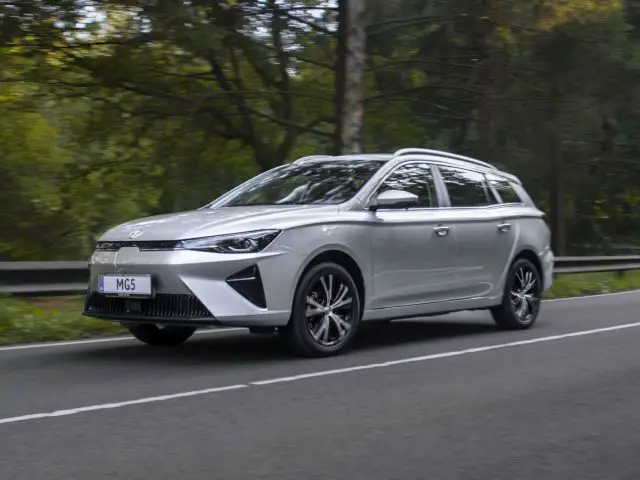 Een slanke zilveren MG5 Electric stationwagen rijdt op een bochtige weg met bomen op de achtergrond.