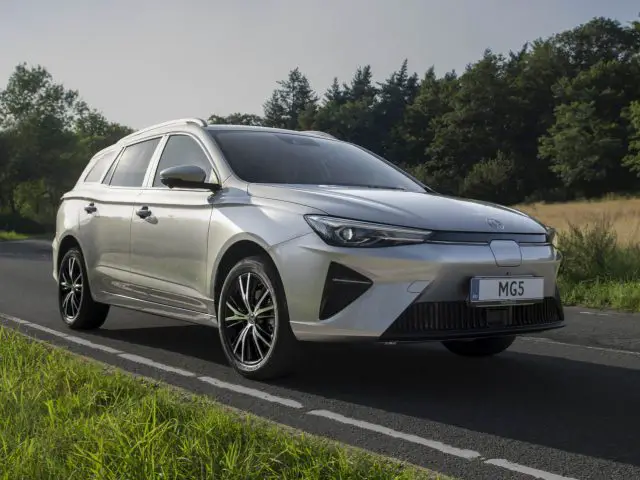 Een slanke MG5 Electric staat geparkeerd op een landelijke weg, omgeven door weelderige groene velden en bomen.