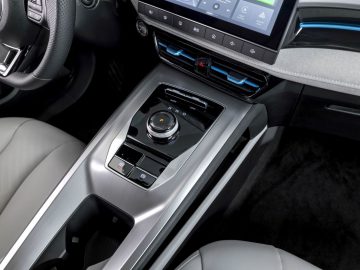 Close-up van de middenconsole van de MG5 Electric met een versnellingspookknop, bekerhouder, klimaatregelingsknoppen en een touchscreen-display erboven. Het interieur heeft een strak design met metallic en lederen afwerkingen.