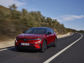 Een rode Renault Megane E-Tech elektrische auto glijdt soepel over een verharde weg, omgeven door weelderige vegetatie onder een helderblauwe lucht.