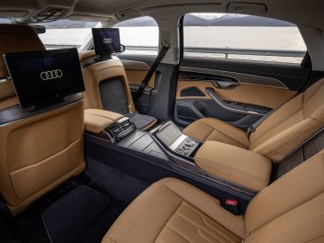 Het interieur van een luxe auto is voorzien van bruinleren stoelen, twee entertainmentschermen op de achterbank en een middenconsole met bedieningspanelen. Gelegen aan een watermassa met bergen op de achtergrond roept het de vraag op: waarom rijdt het kabinet niet met elektrische auto's?