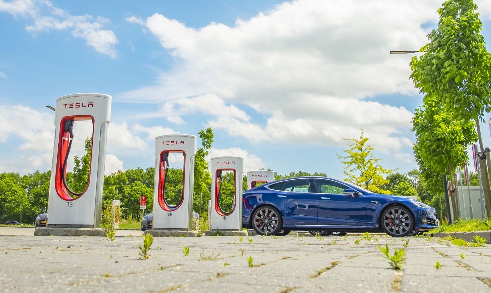 Een strakke blauwe Tesla staat geparkeerd bij een Supercharger-station met vier laadeenheden, omgeven door bomen en een heldere hemel.