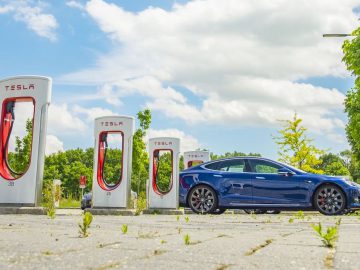 Een blauwe elektrische auto staat geparkeerd naast verschillende Tesla Supercharger-stations onder een gedeeltelijk bewolkte hemel in een groen aangelegd gebied, wat de naadloze mix van milieuvriendelijke technologie en natuur benadrukt.