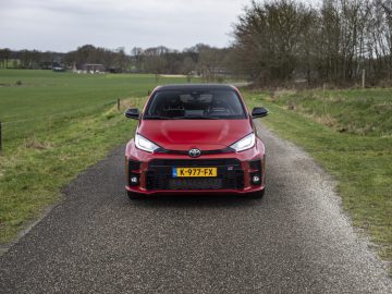 Een rode Toyota GR Yaris met kenteken "K-977-FX" staat geparkeerd op een smalle landelijke weg omgeven door groene velden en bomen.