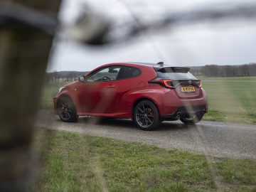 Een rode Toyota GR Yaris staat geparkeerd op een landelijke weg, omgeven door groene velden en bomen, met vage takken op de voorgrond.