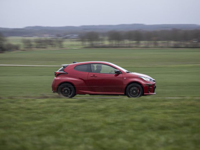 Een rode Toyota GR Yaris rijdt snel over een landelijke weg met groene velden en bomen op de achtergrond.